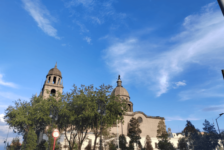 Fotografía de una iglesia con una cúpula y una torre, rodeada de árboles y faroles, bajo un cielo azul con algunas nubes.